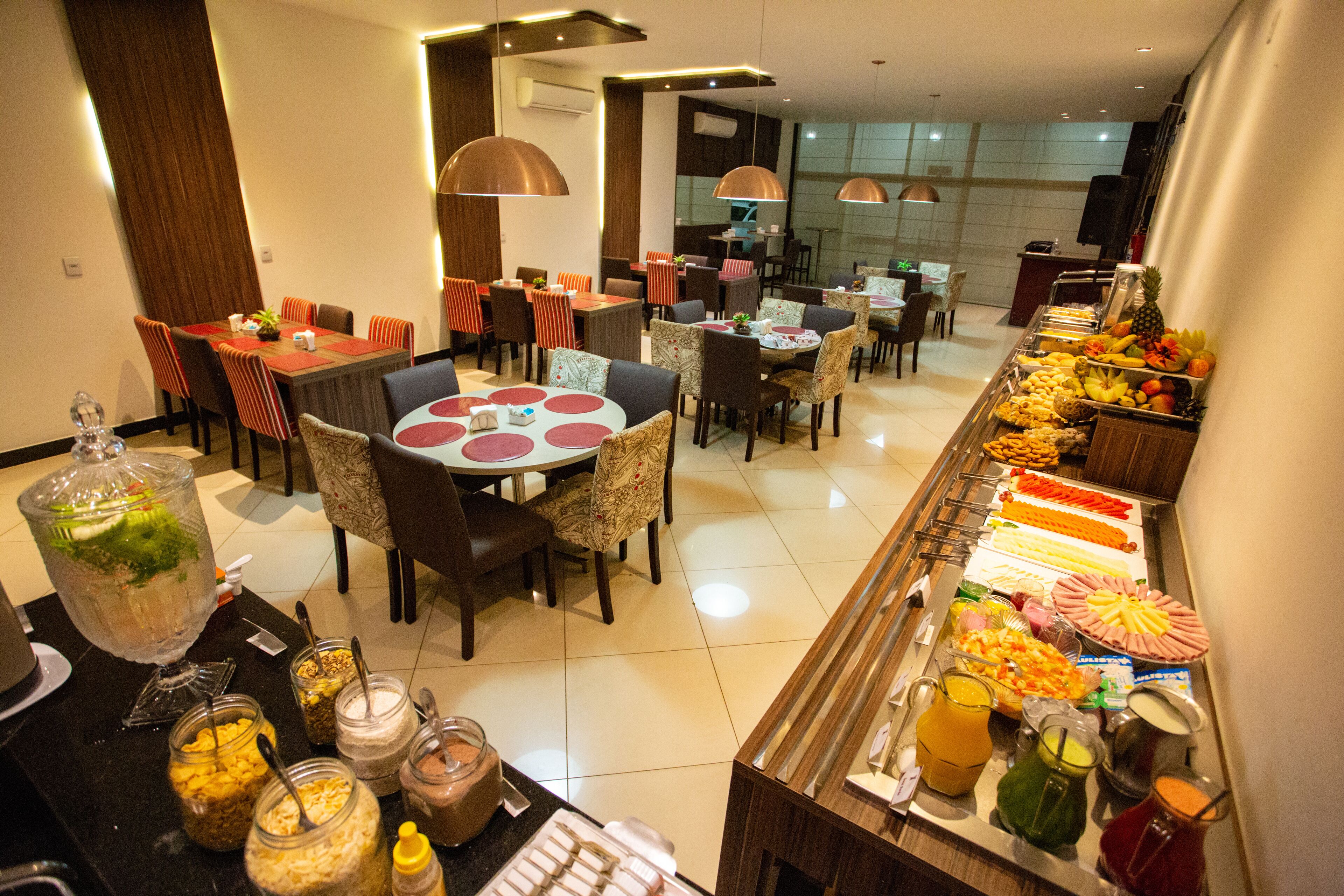 A hotel dining area with multiple tables and chairs, a buffet with various food items, and a fruit display.