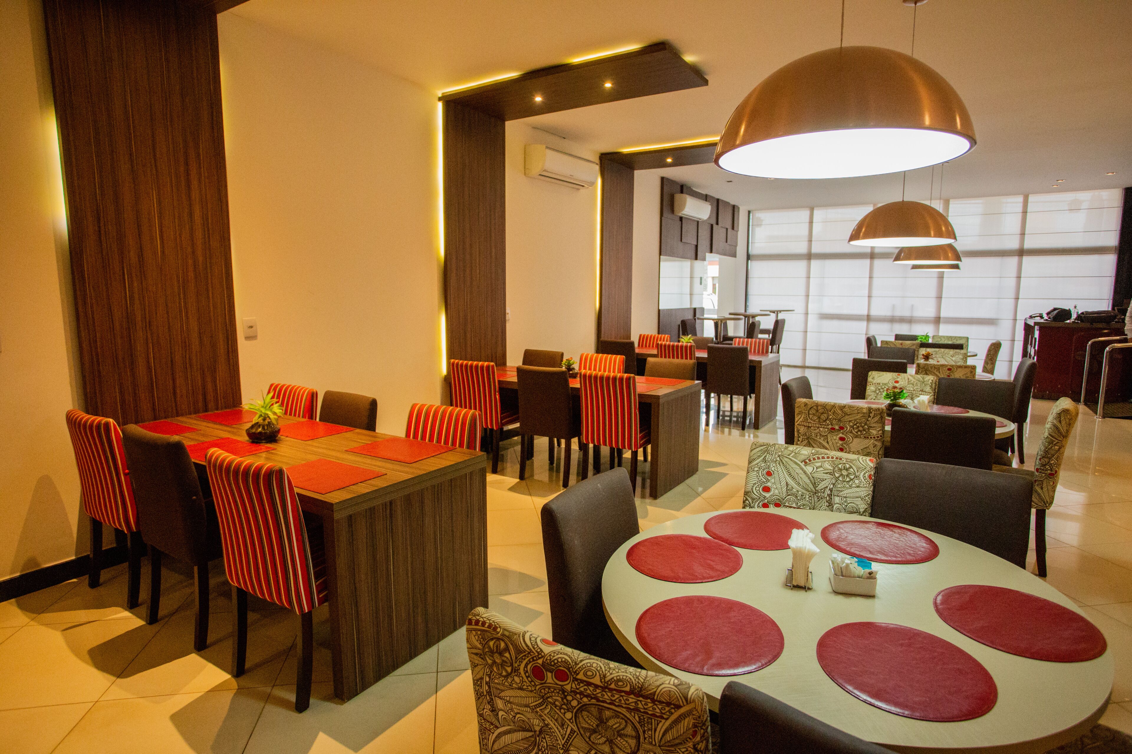 Un restaurante moderno con varias zonas de comedor, incluyendo una mesa central con individuales rojos y una lámpara colgante grande encima.