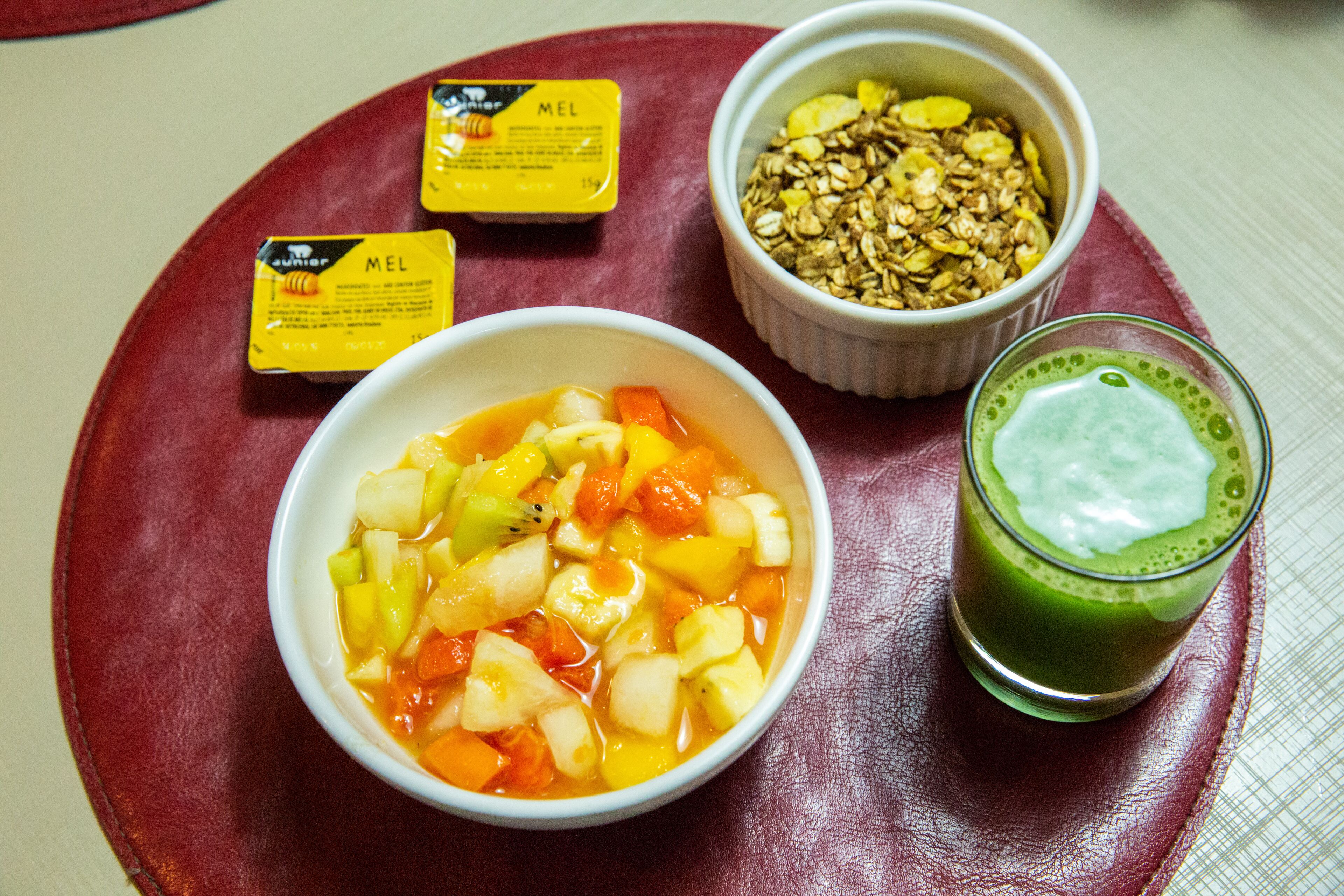 A meal with a bowl of fruit salad, a bowl of granola, a glass of green smoothie, and two packets of honey.