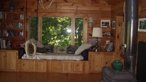 Interior acogedor de una cabaña con paredes de madera, un ventanal con vista a los árboles, un sofá con cojines, una lámpara y un estufa de leña.