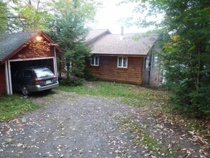 Eine Garage mit einem geparkten Auto, ein Holthaus mit Schornstein und ein Schotterweg, umgeben von Bäumen.