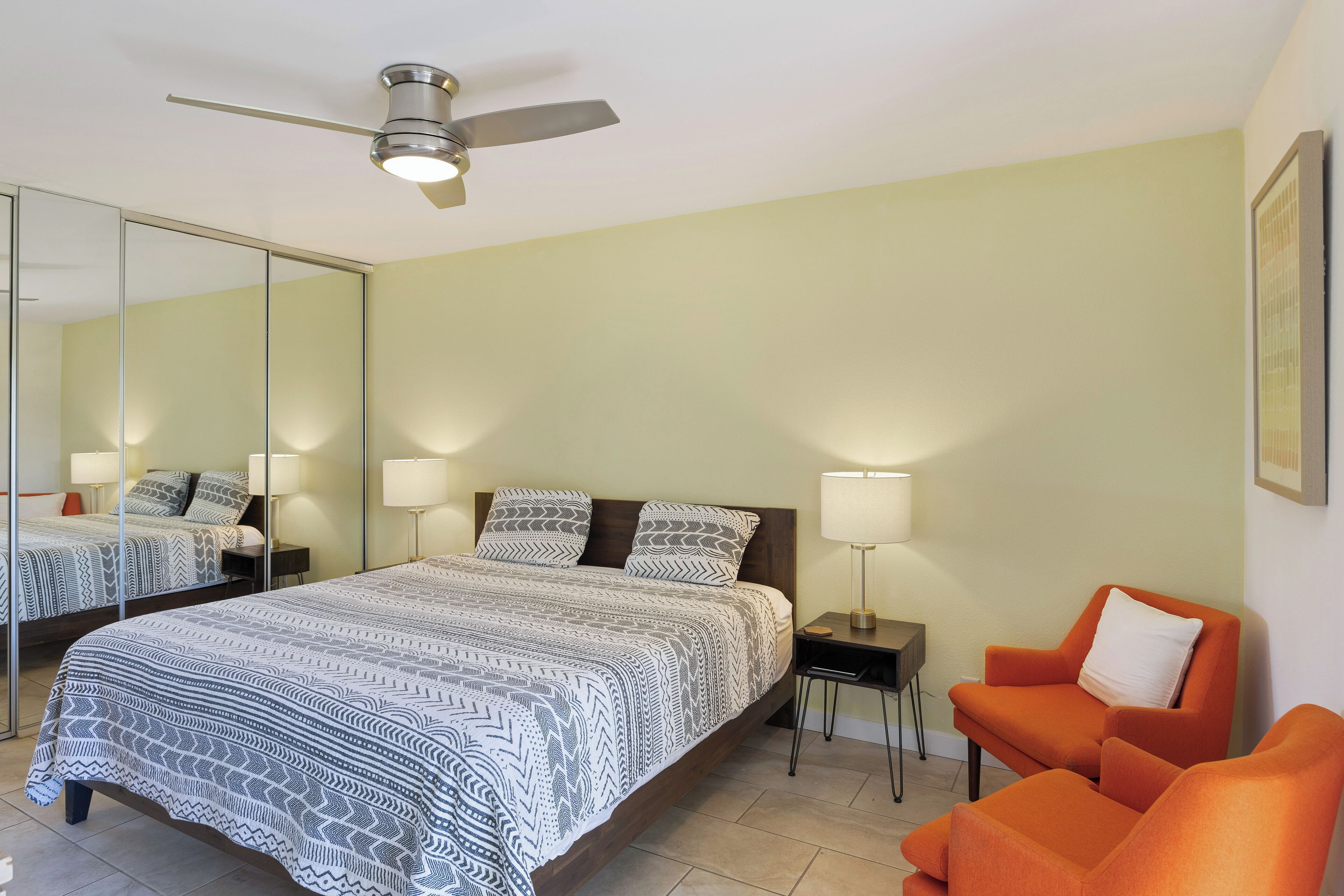 A bedroom with a large bed, a nightstand, a chair, a ceiling fan, and a mirror on the wall.