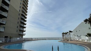 A swimming pool surrounded by a brick walkway, with a high-rise building on one side and a palm tree-lined area on the other.