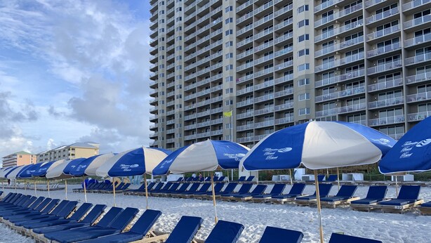 A beach with rows of blue lounge chairs and umbrellas, a tall building in the background, and a sandy beach.