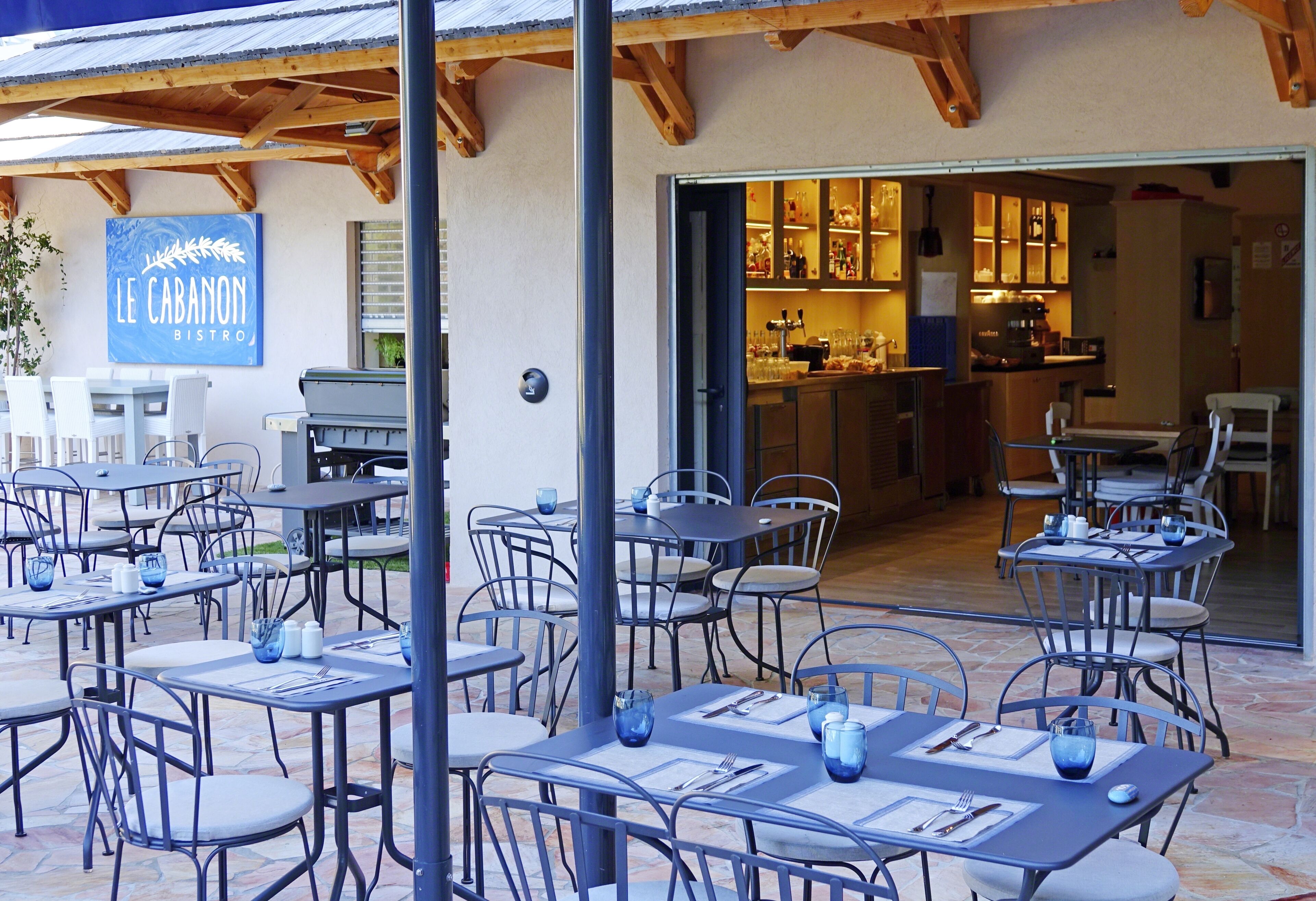 Un café en plein air avec des tables et des chaises, une enseigne indiquant « Le Cabanon Bistro », et une cuisine visible à travers une porte ouverte.