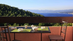 Uma mesa posta com xícaras de chá brancas e uma chaleira em uma varanda com vista panorâmica para um corpo d'água e montanhas ao longe.