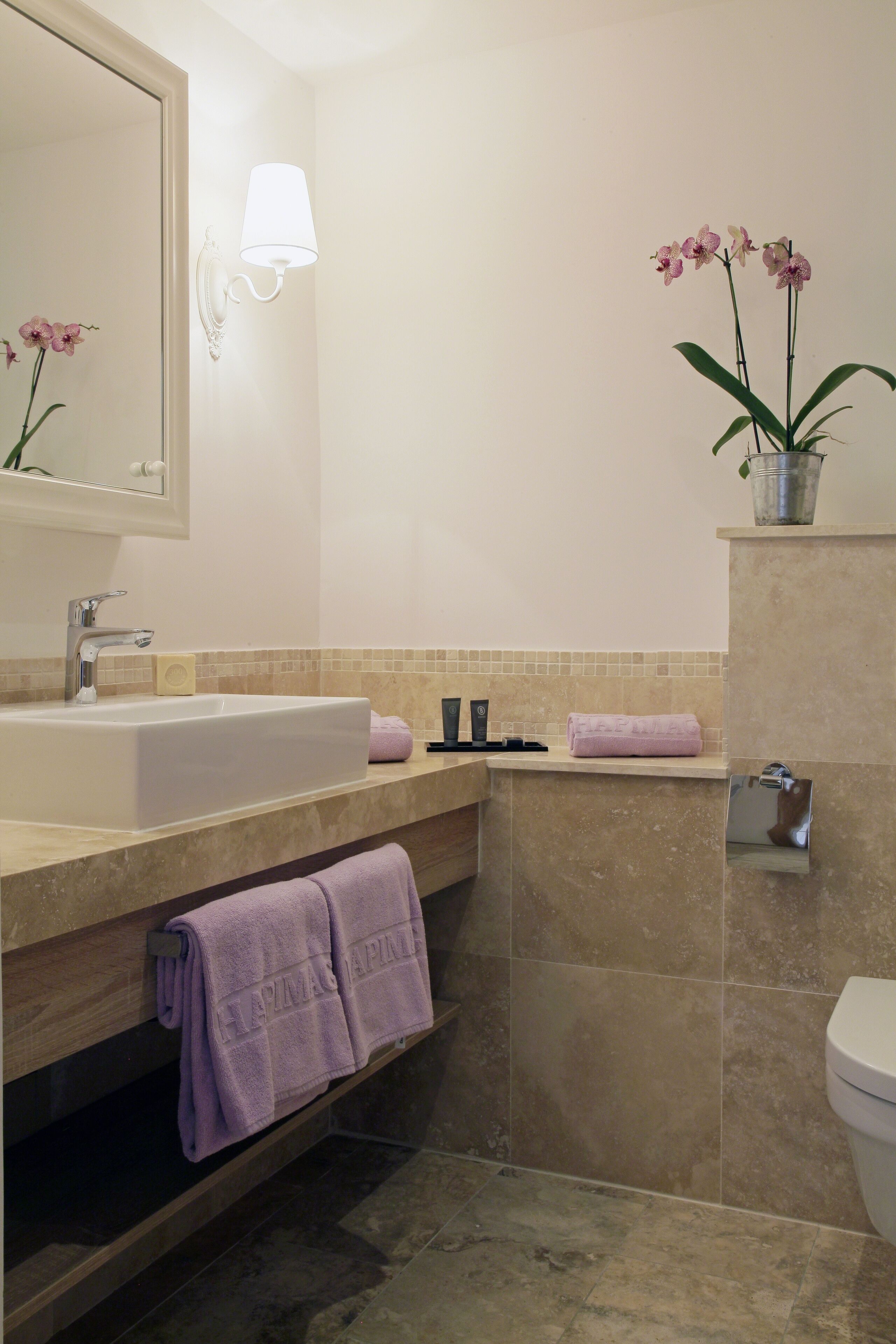 Ein Badezimmer mit Waschbecken, Spiegel und Toilette. Handtücher und ein Topf mit einer Orchidee stehen auf der Ablagefläche.