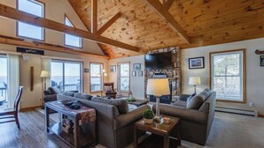 A cozy living room with wooden beams, a stone fireplace, and large windows offering views. - Serenity Place: 5 bedroom lake home, hot tub, pool table/game room (Penn Yan)
