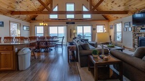 A spacious living area with a wooden ceiling, a dining area with a table and chairs, a sofa, and a stone fireplace. - Serenity Place: 5 bedroom lake home, hot tub, pool table/game room (Penn Yan)