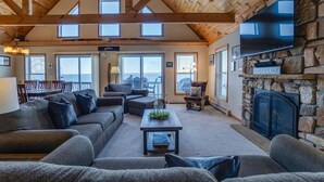 A spacious living room with a stone fireplace, large windows, and a high ceiling with exposed beams. - Serenity Place: 5 bedroom lake home, hot tub, pool table/game room (Penn Yan)