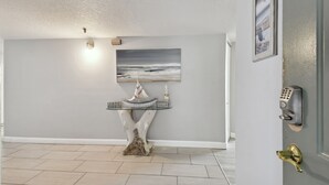 Un pasillo de diseño moderno, con un cuadro colgado en la pared que muestra una escena de playa, una mesa decorativa con un velero y una puerta con cerradura electrónica.