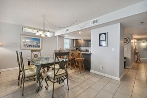 A spacious kitchen with modern appliances, a dining area with a glass table and metal chairs, and a living area with a large clock on the wall.