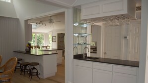 A kitchen with white cabinetry, a black countertop, and a glass wine rack. There are wicker chairs and a small island with stools. - Sea Pines Home with Sparkling Heated Pool-Hole #2 Harbour Town Golf Links-Grill (Hilton Head Island)