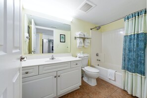 A bathroom with a white vanity, a toilet, a shower with a blue curtain, and a towel rack with folded towels. - Amazing Oceanfront Condo, Outdoor Pool and Hot Tub | Crescent Keyes - 1006 (North Myrtle Beach)
