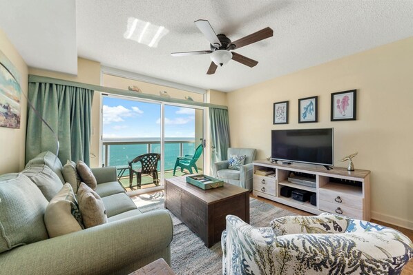 A living room with a sofa, armchair, coffee table, TV stand, and artwork on the wall. - Amazing Oceanfront Condo, Outdoor Pool and Hot Tub | Crescent Keyes - 1006 (North Myrtle Beach)