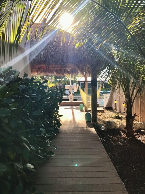 A wooden pathway leading to a thatched-roof structure, surrounded by lush greenery and palm trees.