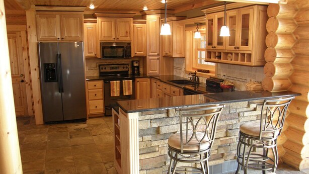A spacious kitchen with wooden cabinets, stainless steel appliances, a large island with bar stools, and a stone accent wall. - Luxurious Log Home with Mille Lacs Lake Sunrise Views (Garrison)