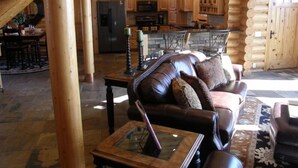 A log cabin interior with a living room featuring a leather sofa, a glass-top coffee table, and a patterned rug. The kitchen area is visible in the background with wooden cabinets and a stone countertop. - Luxurious Log Home with Mille Lacs Lake Sunrise Views (Garrison)