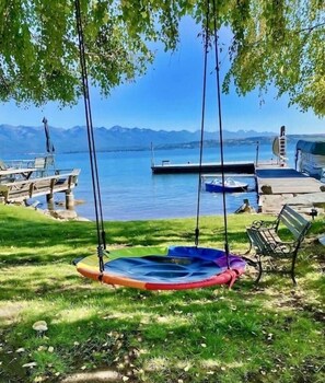 Une zone herbeuse avec une aire de jeux colorée comprenant des balançoires, une jetée en bois et un bateau sur un lac, avec des montagnes en arrière-plan.