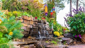 Un jardín con una pequeña cascada, casas para pájaros de colores y abundante vegetación.
