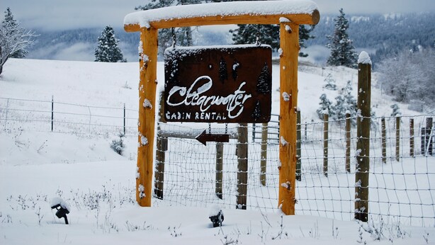 A wooden cabin rental sign with a directional arrow, surrounded by a snow-covered landscape and a fence. - Peaceful, Private Cabin overlooking the Clearwater River. Dbl occ rates (Orofino)