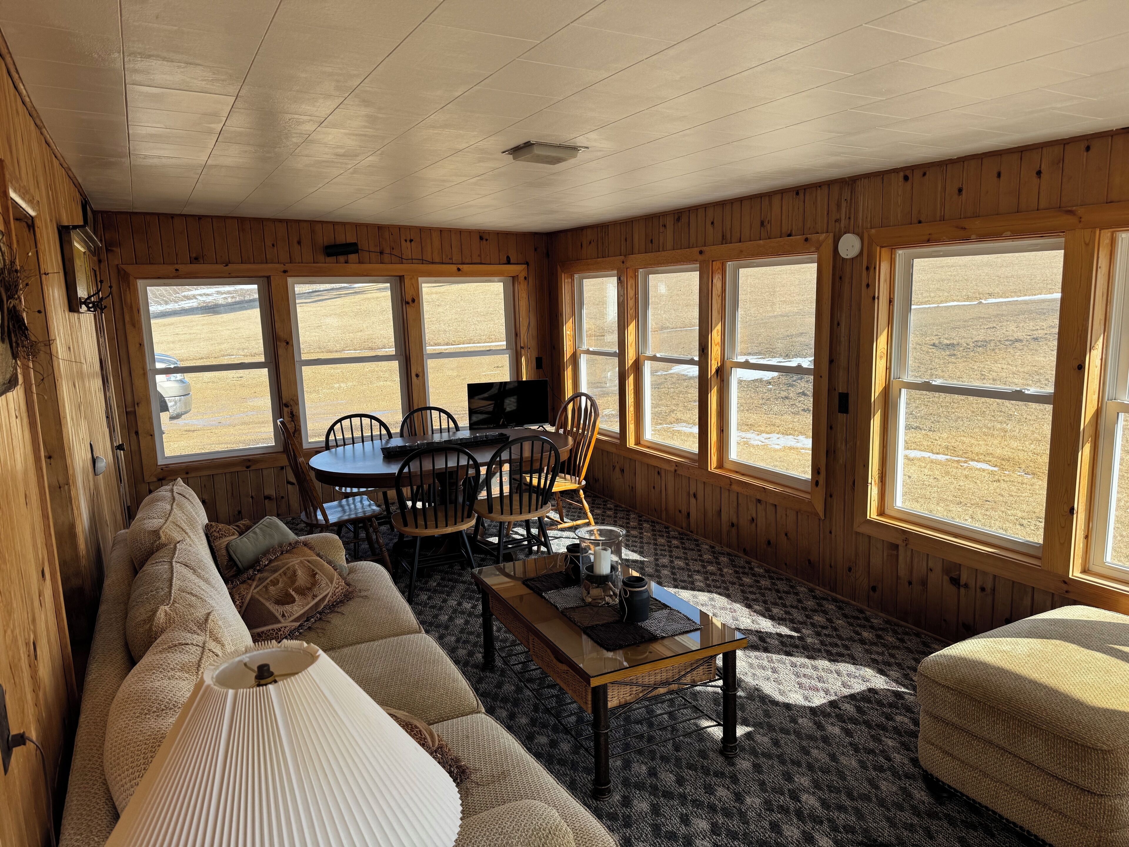 L’intérieur chaleureux d’un chalet, avec un décor en bois, un canapé, une table à manger et des chaises, ainsi que de grandes fenêtres donnant sur un paysage enneigé.