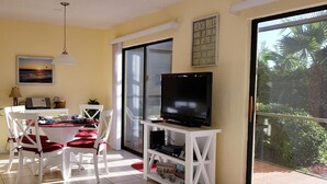Une pièce lumineuse, avec une table à manger, des chaises, un téléviseur posé sur un meuble, une plante et une grande fenêtre donnant sur un paysage verdoyant.