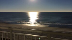 En vy över stranden med solen som speglar sig i vattnet, ett räcke i förgrunden och en klar himmel.