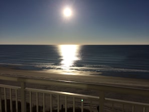 Une vue sur la plage, avec le soleil se reflétant sur l’eau, une rambarde au premier plan et un ciel dégagé.