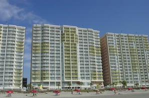 Un hôtel de grande hauteur doté de nombreux balcons, situé en bord de mer, et dont le rez-de-chaussée est agrémenté de parasols rouges.
