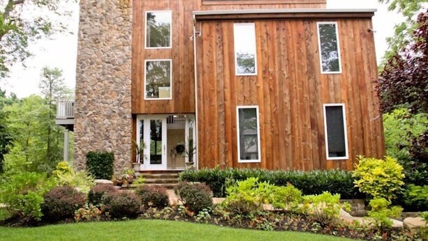 A modern two-story house with a stone facade and wooden paneling, surrounded by well-maintained landscaping.
