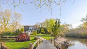 A wooden bridge leading to a house with a garden and a pond.