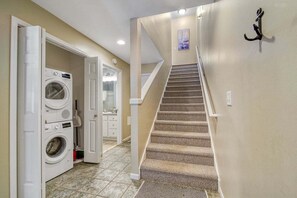 Une buanderie équipée d’un lave-linge et d’un sèche-linge, un escalier dont les marches sont recouvertes de moquette, et une porte donnant sur une autre pièce.