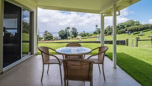 Une terrasse couverte, dotée d’une table ronde et de quatre chaises en osier, qui offre une vue sur une pelouse bien entretenue et sur l’océan.