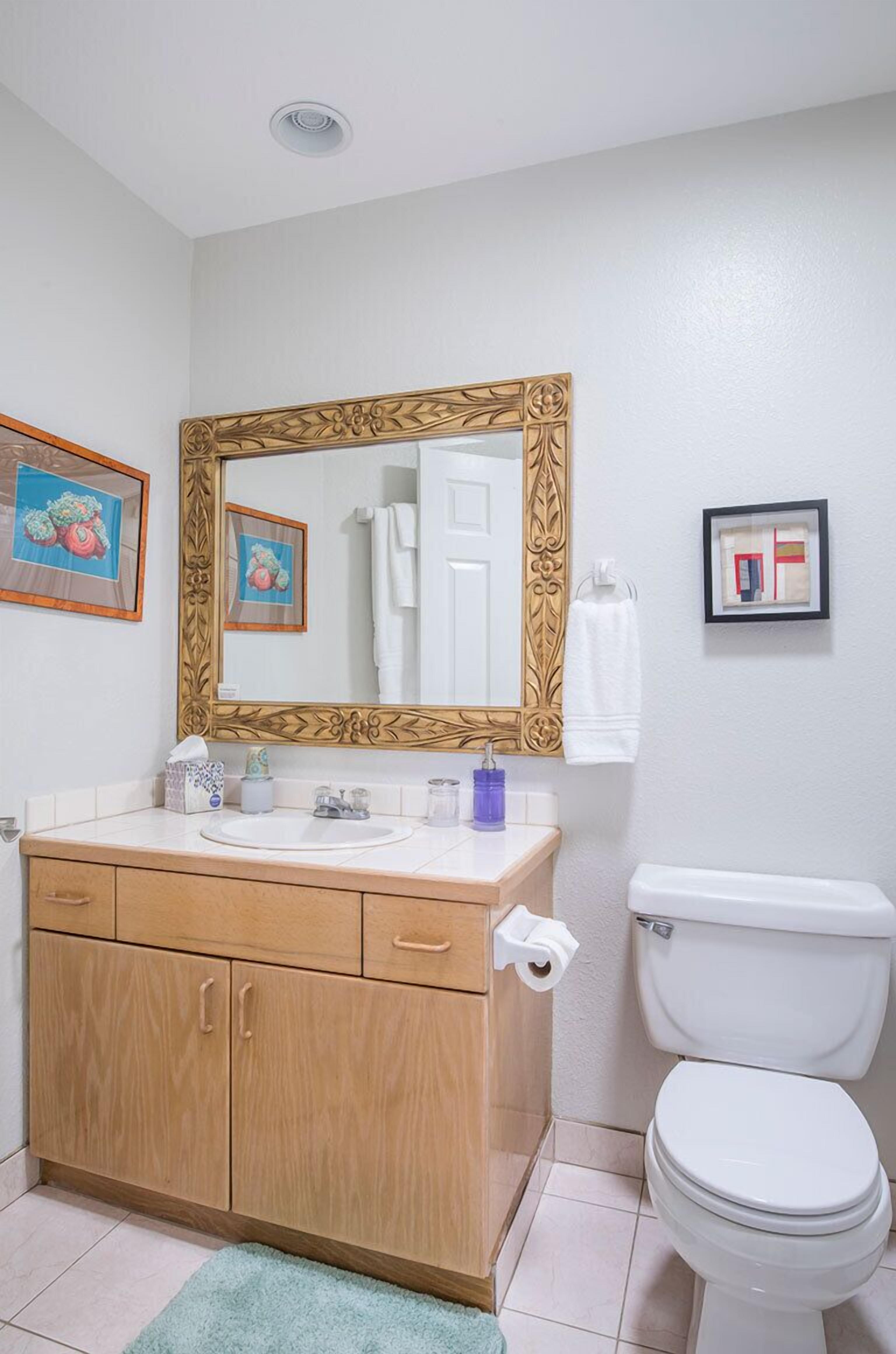 A bathroom with a wooden vanity, a large mirror with an ornate frame, a toilet, and a small framed picture on the wall.