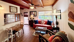 A cozy living room with a wooden ceiling, a red sofa, a dining table with chairs, and a kitchen area in the background.