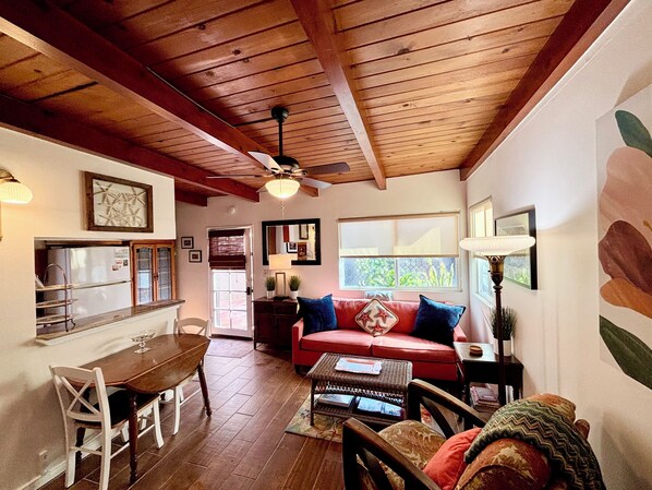 A cozy living room with a wooden ceiling, a red sofa, a dining table with chairs, and a kitchen area in the background.