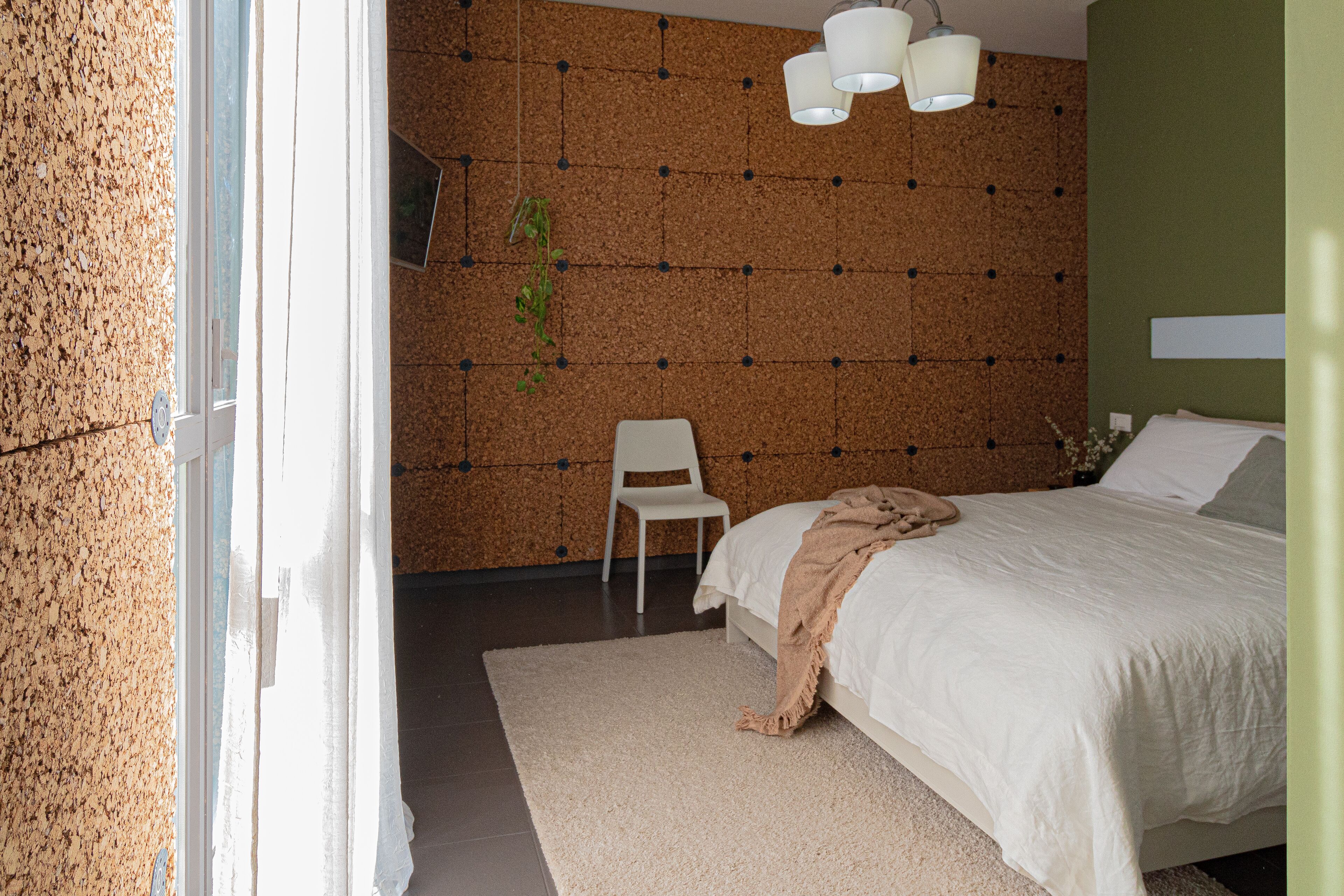 Un dormitorio con pared de corcho, una cama con ropa de cama blanca, una silla blanca y una planta colgante.
