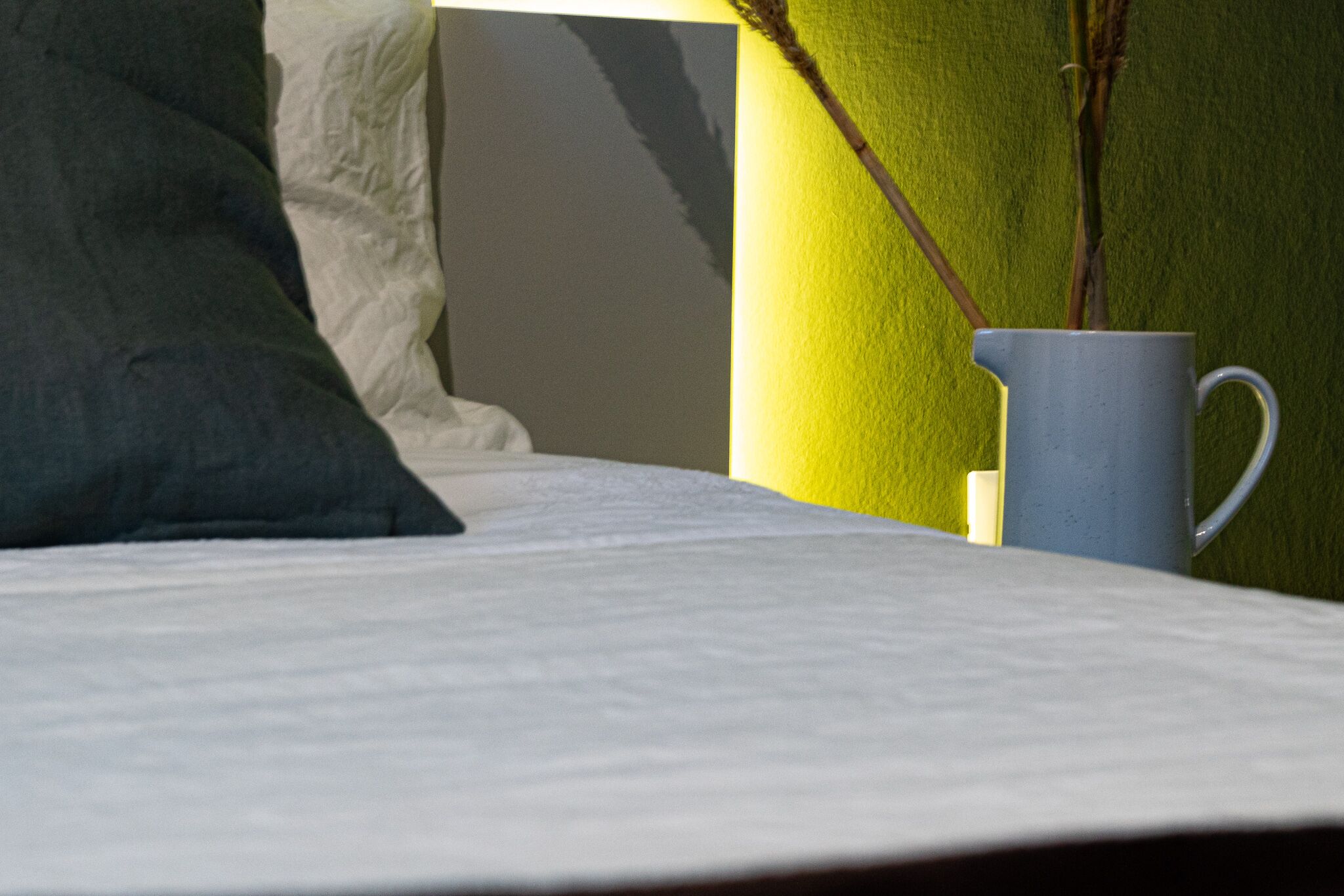A bed with a pillow, a white wall, and a blue mug on a table.