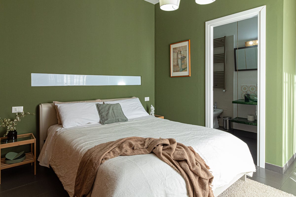 A bedroom with a bed, a nightstand, a framed picture, and a bathroom visible through an open door.