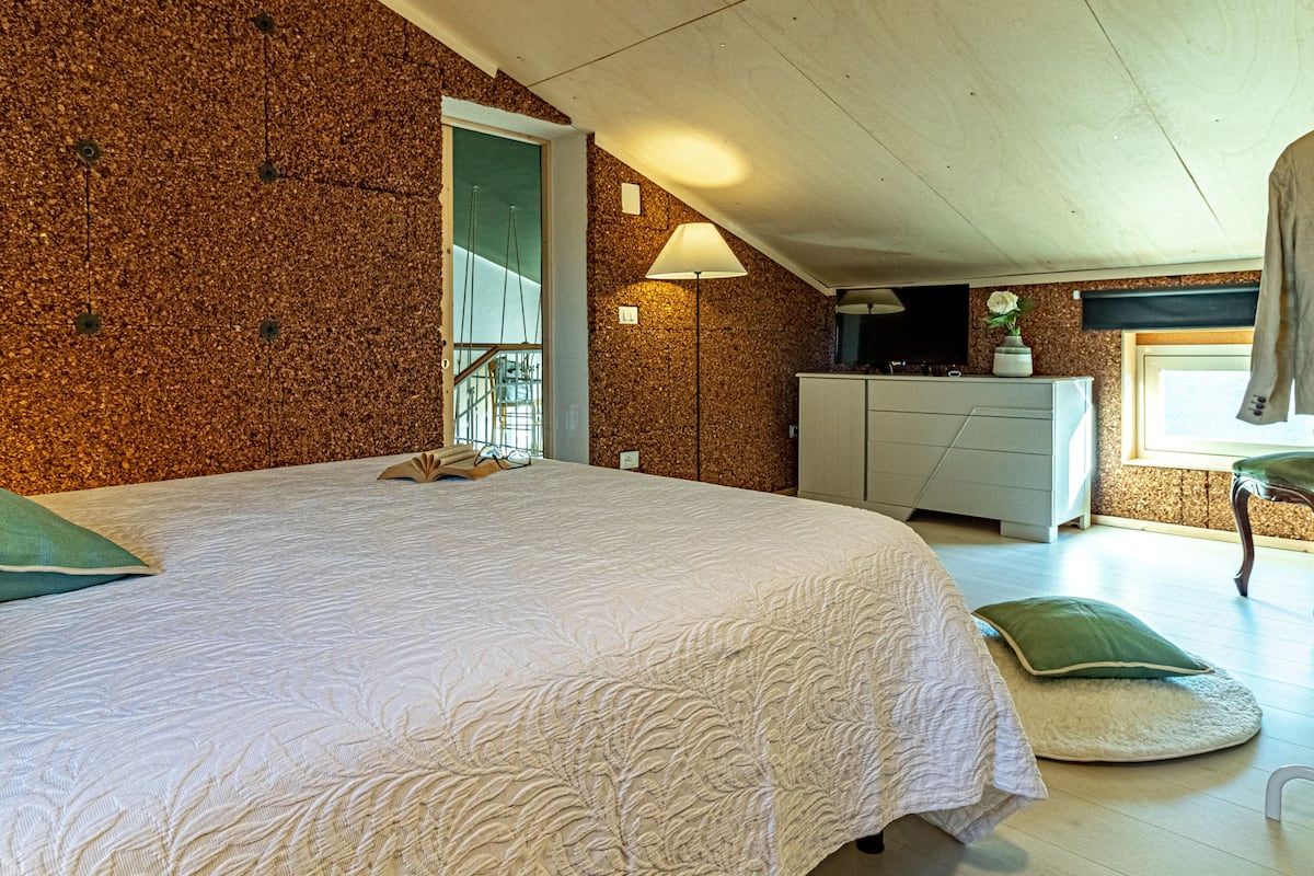 A bedroom with a large bed, a dresser, a window with a view, and a ceiling with exposed beams.