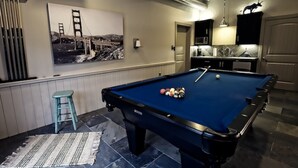 Une table de billard avec des billes, une table de billard bleue, un tableau représentant le pont du Golden Gate, un coin cuisine et un tabouret.