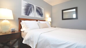 Una cama bien tendida con sábanas blancas, dos lámparas fijadas a la pared y dos cuadros con hojas de palma sobre la cama.