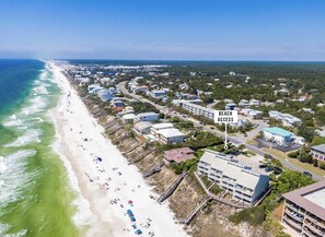 An aerial view of a coastal residential area with beach access, featuring houses, a beach, and a large building with multiple floors.