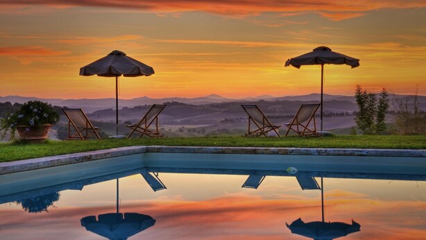 Una piscina con dos sillas de descanso y sombrillas, rodeada de una vista panorámica de colinas y un atardecer.
