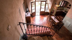 Una escalera conduce a un comedor con una mesa y sillas de madera, una cocina con un lavamanos y armarios, y una vista al exterior a través de una puerta abierta.