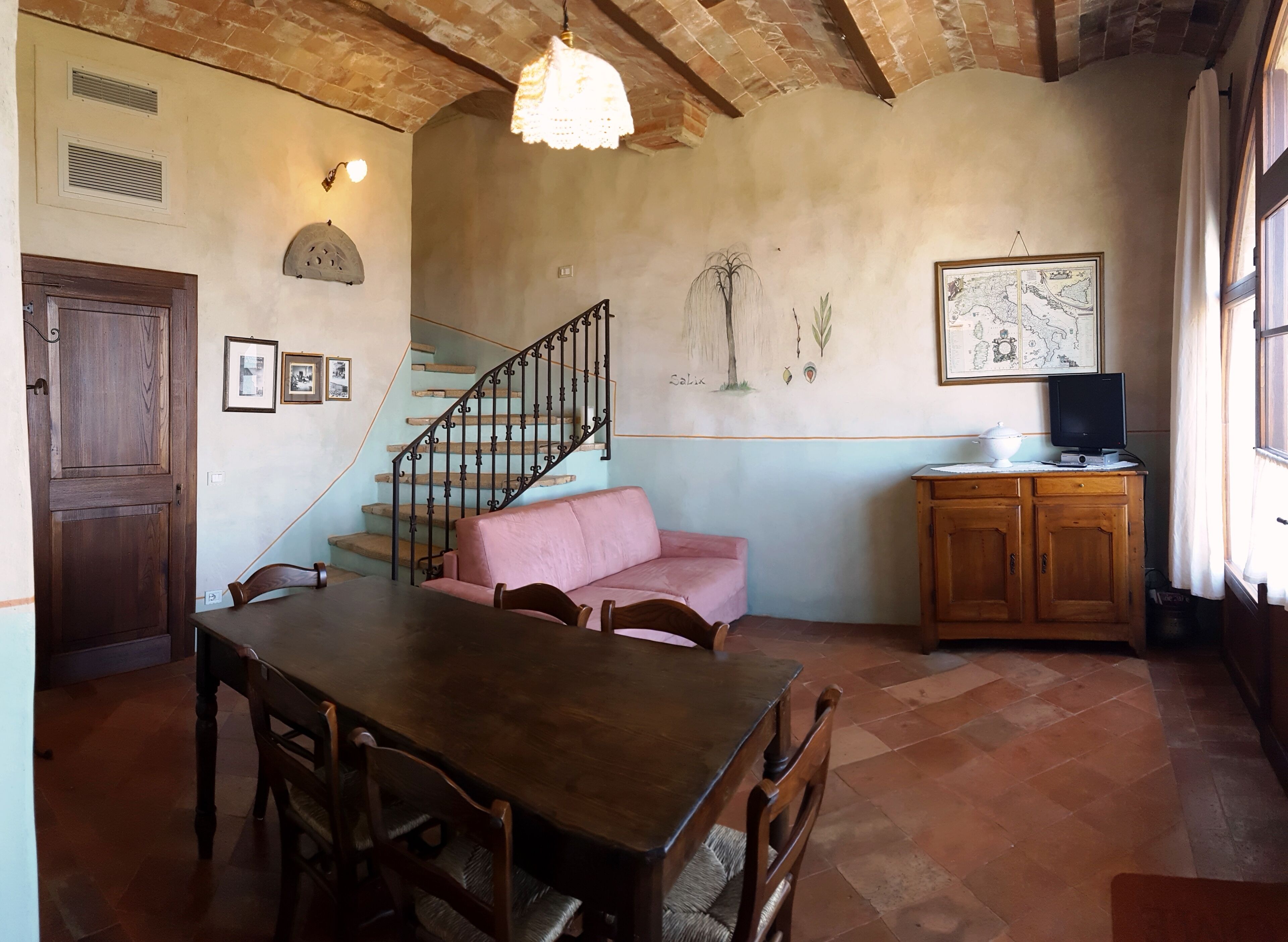 Un comedor con una mesa y sillas de madera, un sofá rosa, una escalera con barandillas negras, un armario de madera y un mapa colgado en la pared.