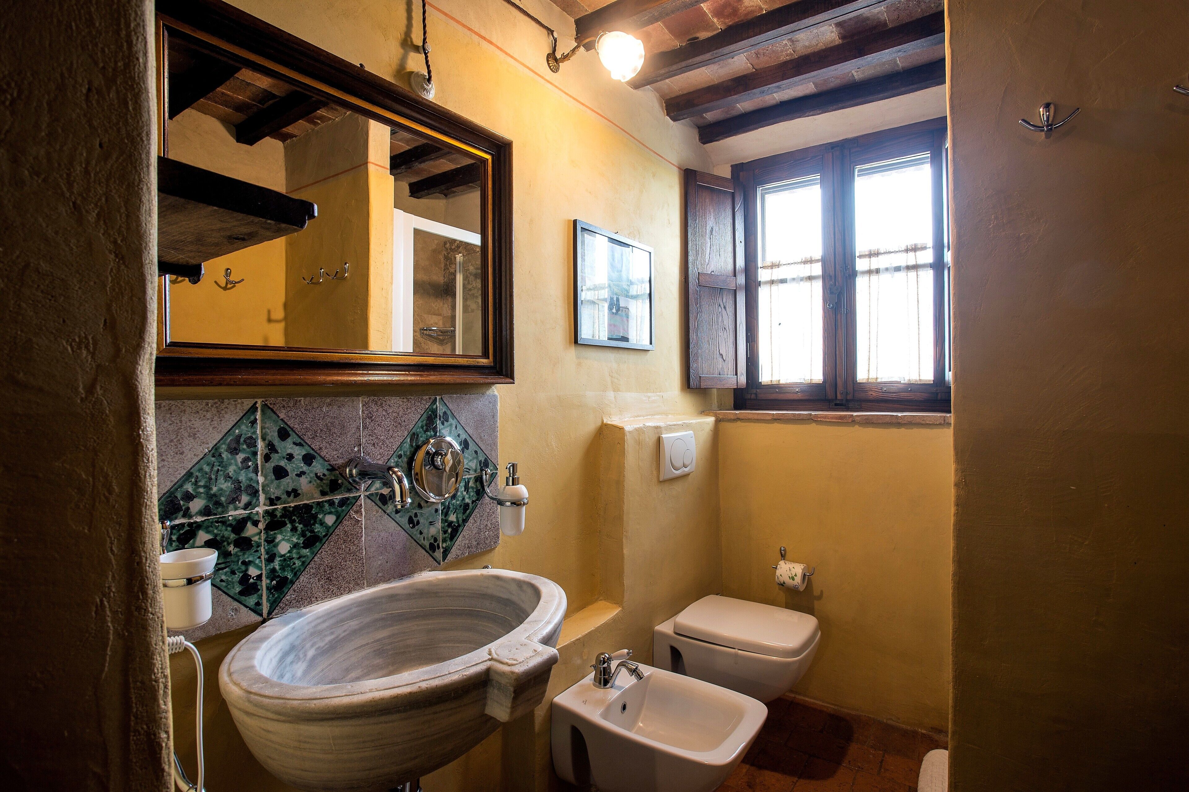 Une salle de bain avec un grand lavabo en céramique, des toilettes et un bidet. Un miroir est placé au-dessus du lavabo et il y a une fenêtre avec des volets en bois.