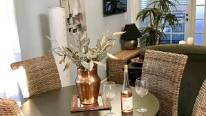 A round dining table with wicker chairs, a copper vase with dried leaves, and a bottle of wine.