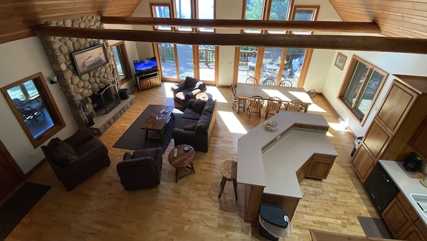 A spacious living area with a high ceiling, wooden beams, a stone fireplace, a large TV, and a view of the outdoors through multiple windows.